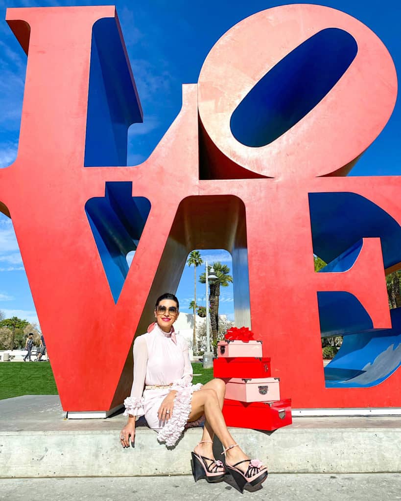 Valentine’s Day Gift Ideas for Her - woman sitting underneath large LOVE sign, in pink dress with pink gift boxes. 