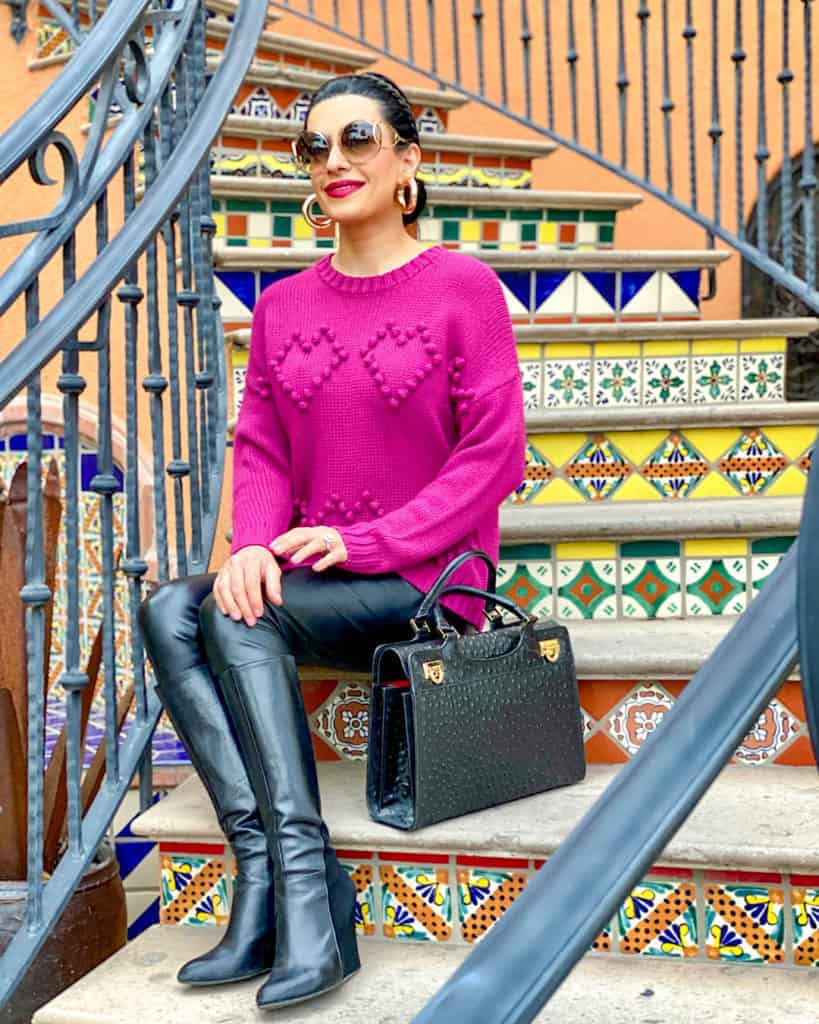 women wearing a pink sweatshirt with hearts, black pants, black boots, and sunglasses sitting on stairs 